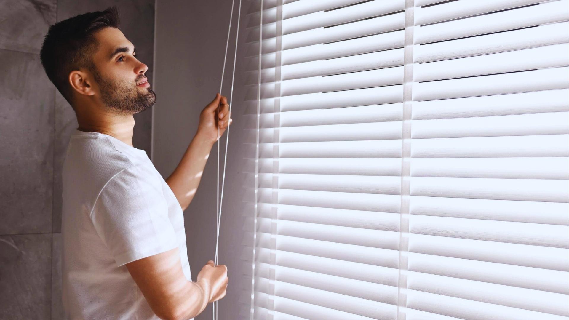 Man in white t-shirt pulls cord to adjust white horizontal blinds on large sunlit window.