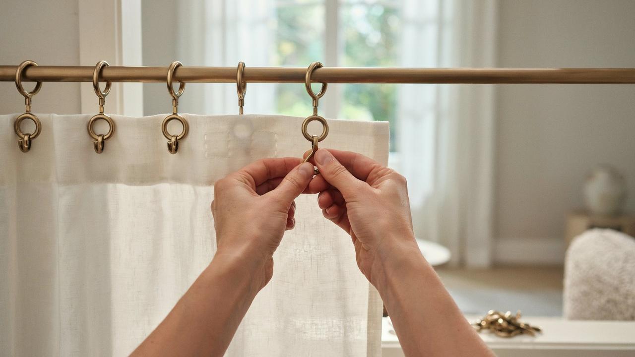 Close-up of hands attaching curtain hooks to decorative rings on a curtain rod.