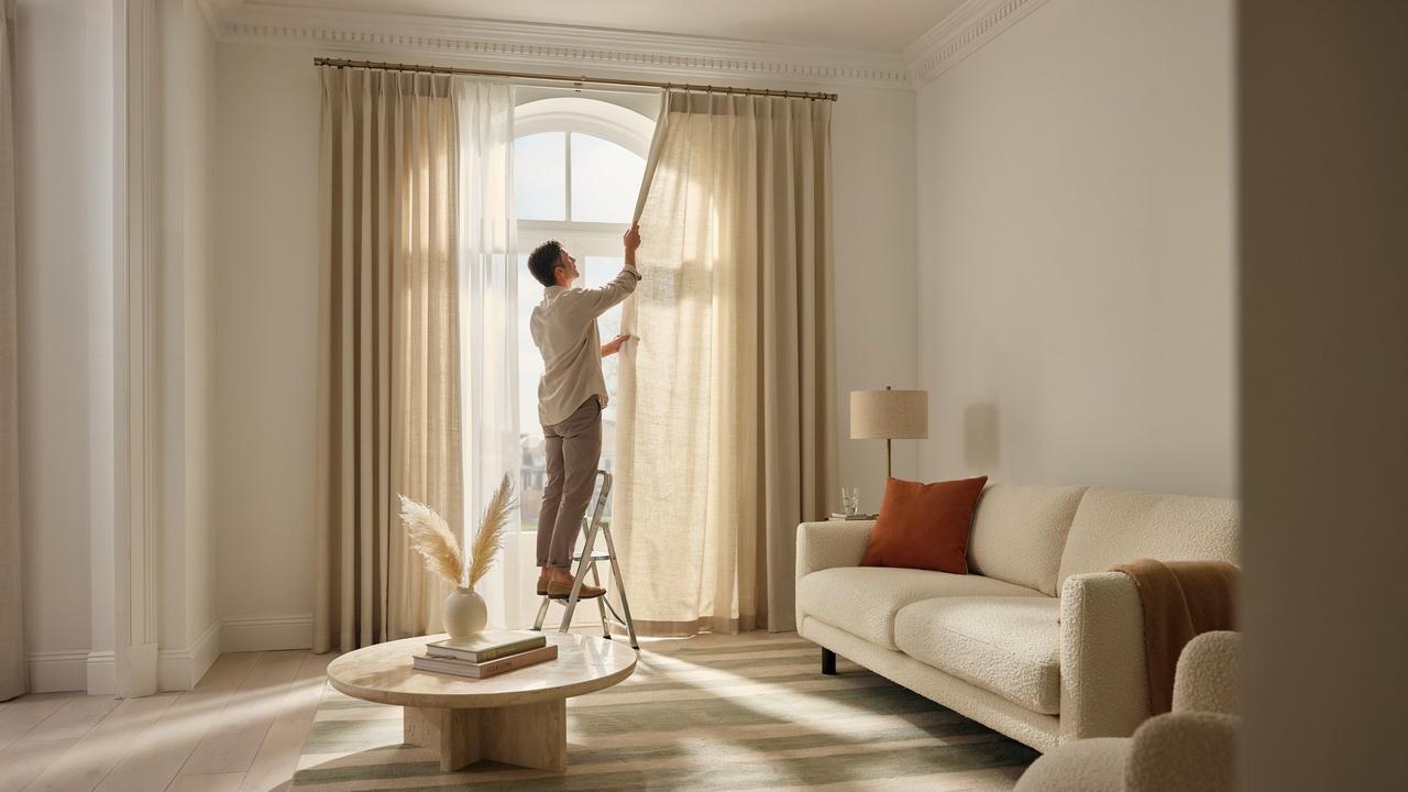 A homeowner installing floor-length curtains beneath decorative crown molding in a modern living room.