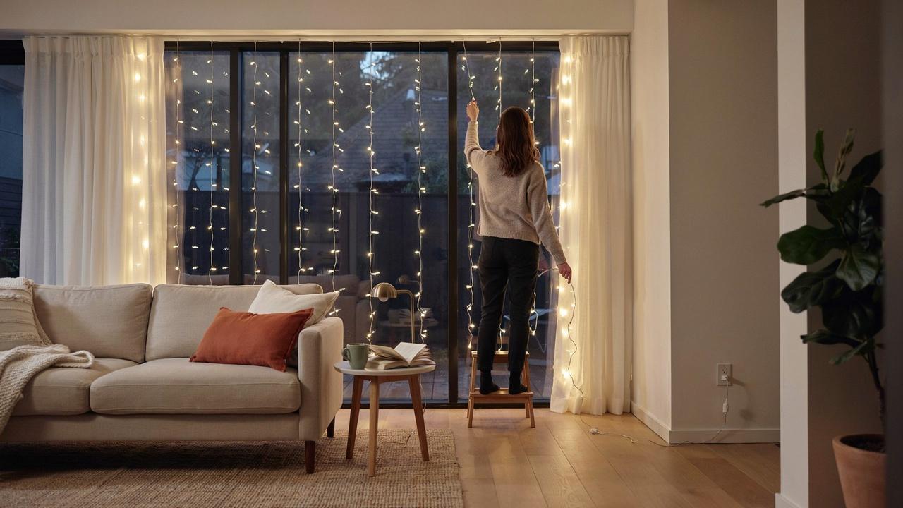 A person securing curtain light wiring and testing the illuminated setup in a cozy room.