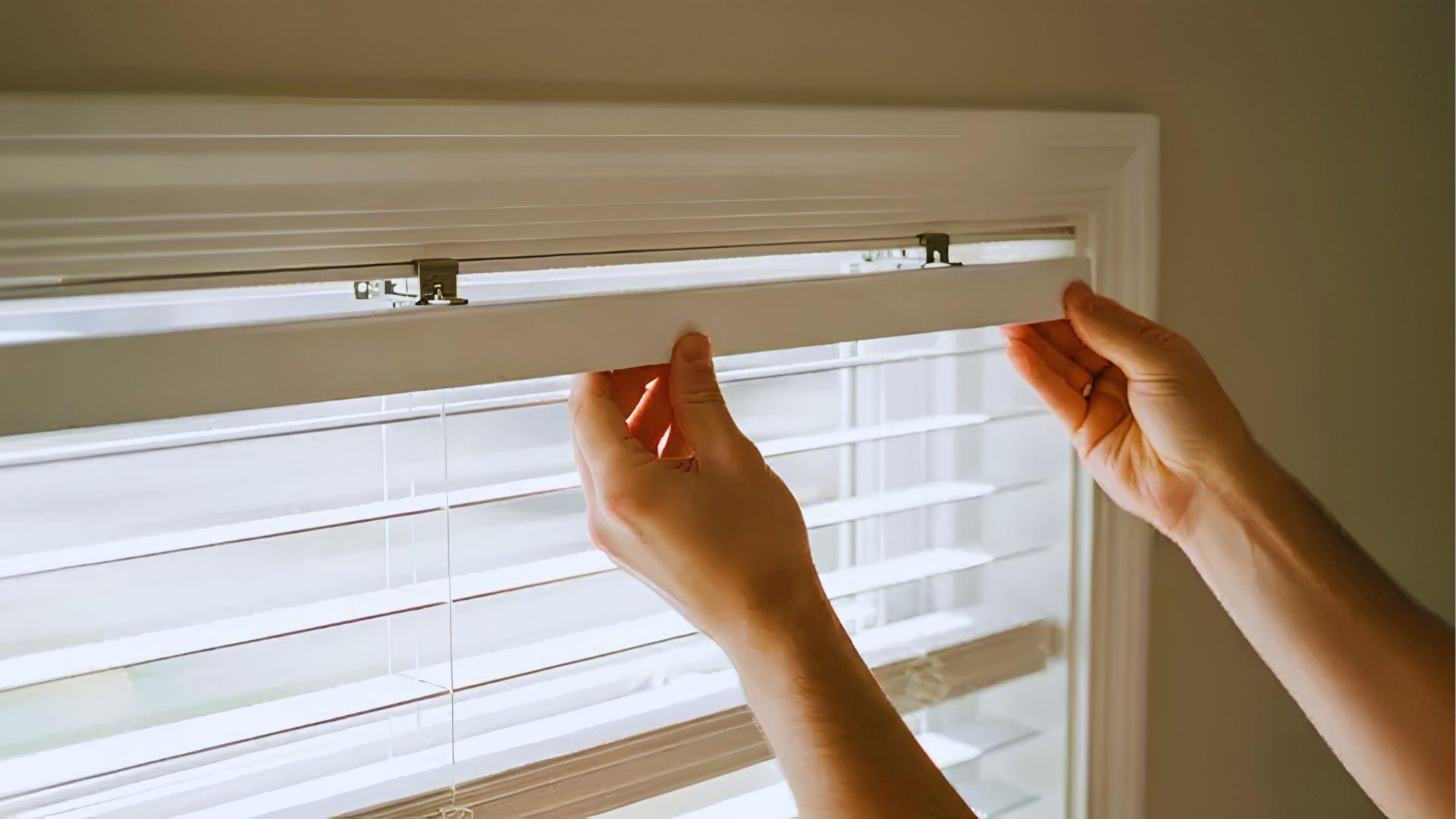 Hands grip bottom rail of white cordless horizontal blinds, lowering them over bright windows.