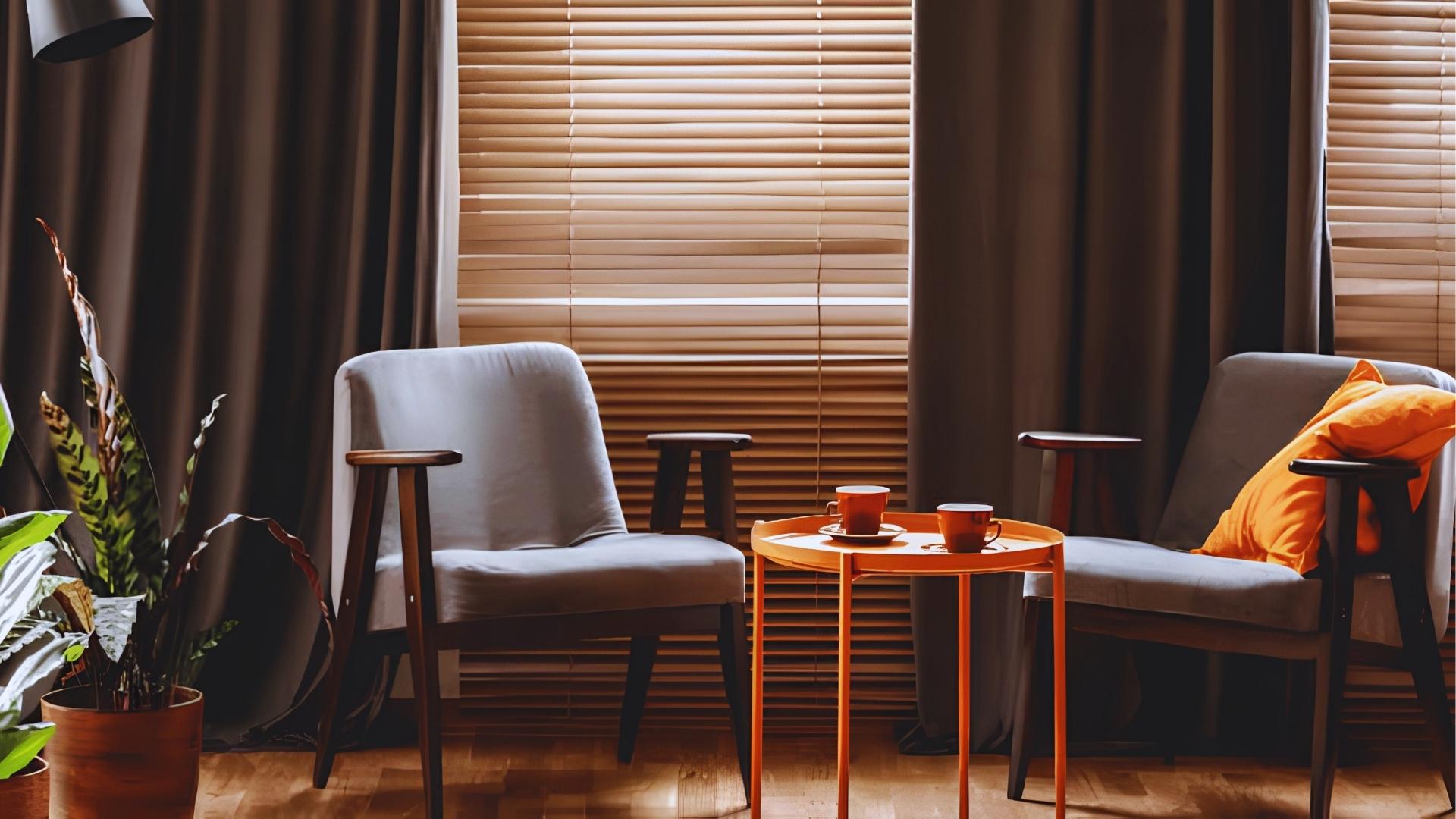 Modern living room with gray armchairs, orange table, closed wooden blinds and dark curtains behind chairs.
