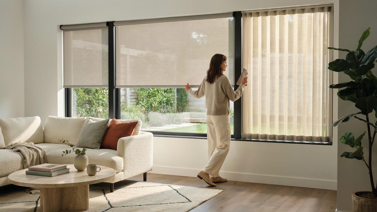 The woman lowers beige solar shade next to sheer cellular shade in a bright living room with sofa.