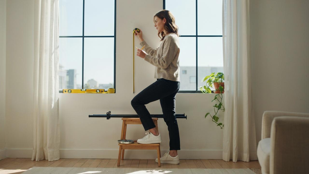 A person measuring and marking the wall above a window to position a curtain rod correctly.