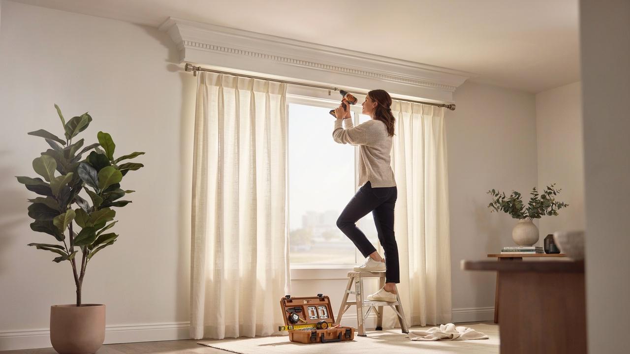 A homeowner installing a curtain rod beneath decorative crown molding in a modern living room.