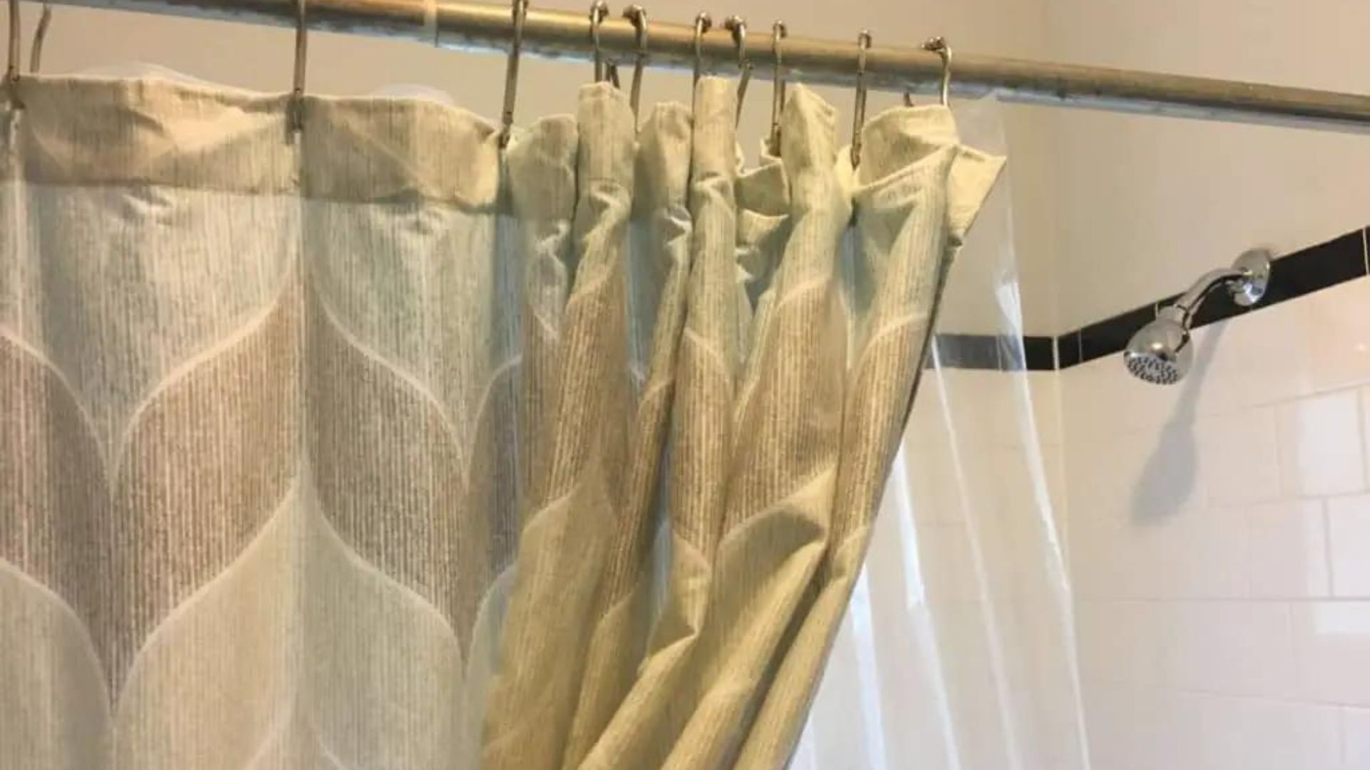 Beige patterned fabric shower curtain on metal rod with hooks, clear liner, chrome showerhead, white subway tile.