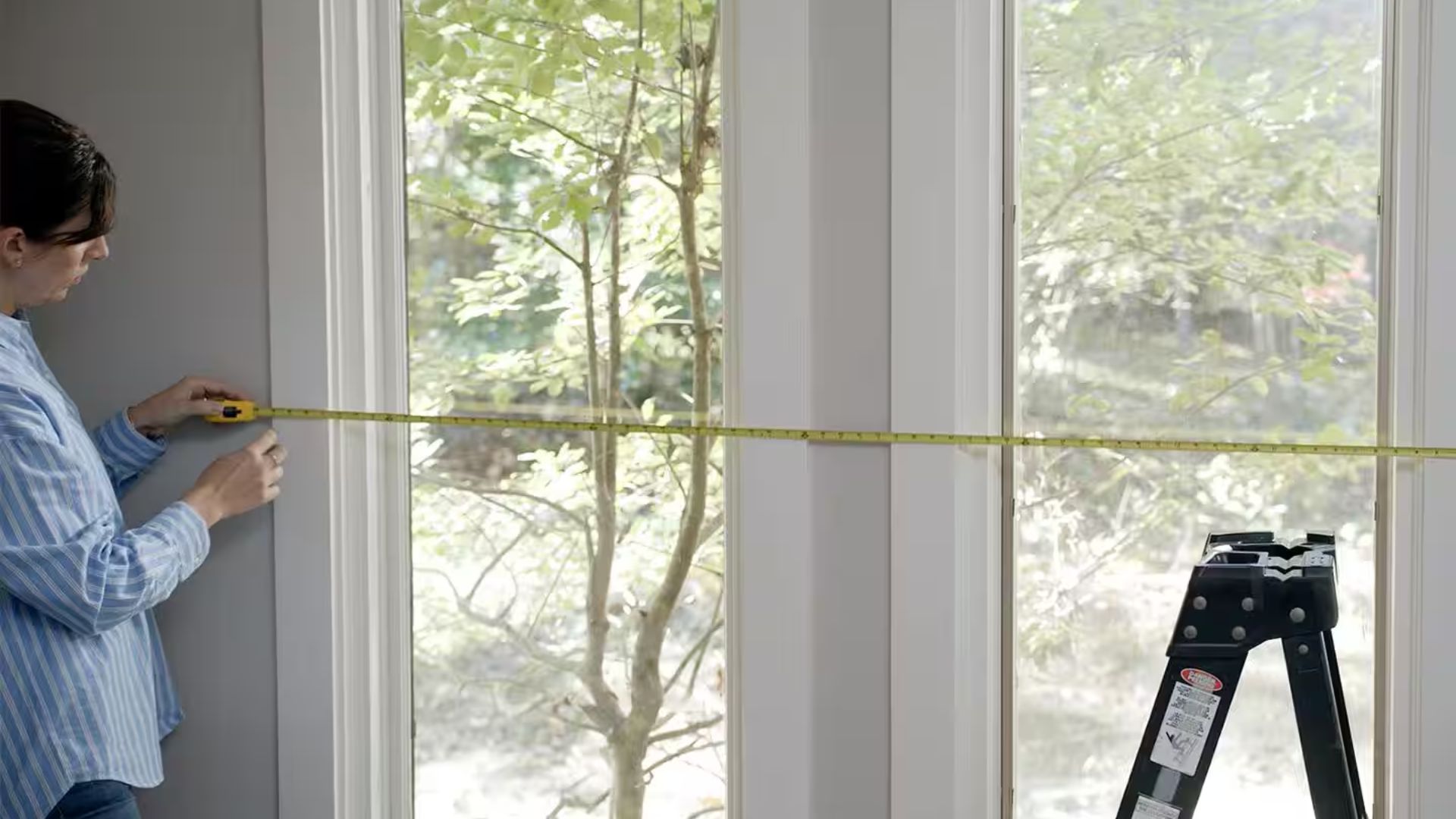 Woman in striped shirt measuring window width with yellow tape measure, ladder nearby, green trees outside.