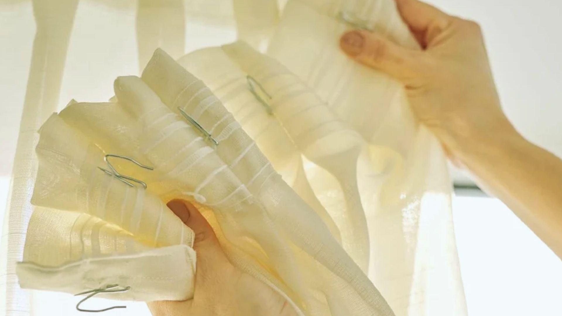 Hands holding white pleated curtain header tape with metal hooks for hanging drapes