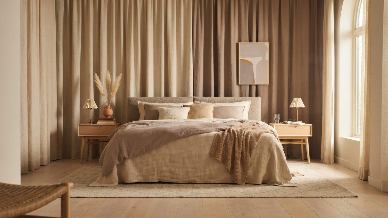 Modern warm bedroom with taupe bedding, beige curtain wall backdrop, arched window, wood nightstands with lamps.
