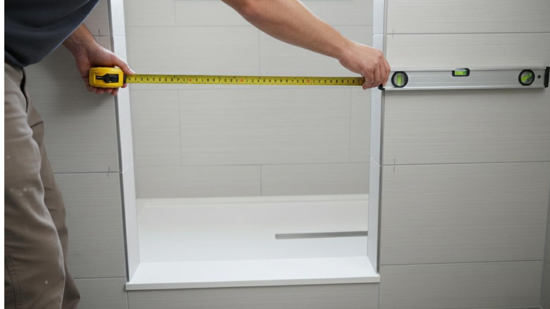Person measuring shower stall width with yellow tape measure, spirit level on gray tiled wall.