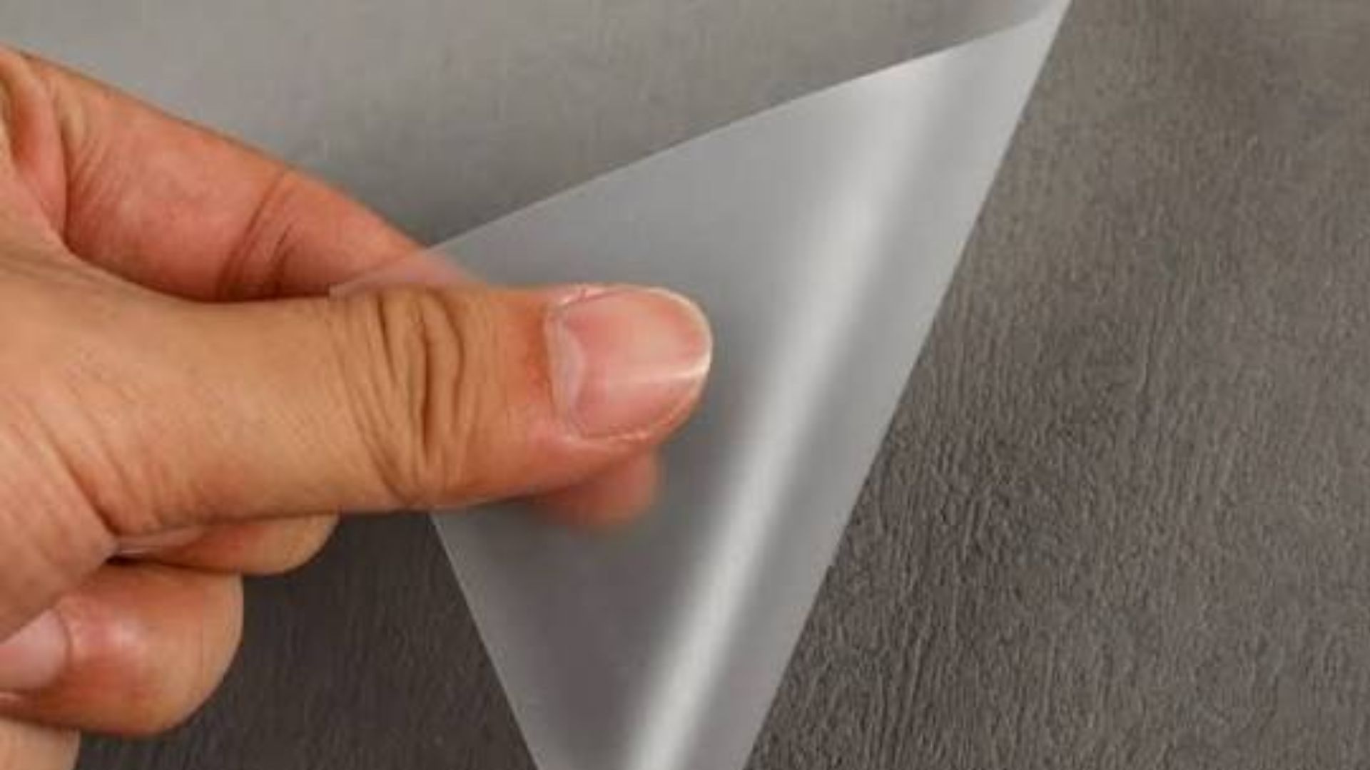 Hand peeling back a translucent frosted protective film from a dark textured surface.